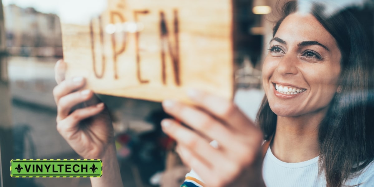 Smiling business owner placing an 'Open' sign on the door, symbolizing a thriving business supported by reliable commercial roofing solutions, featuring the Vinyltech logo.