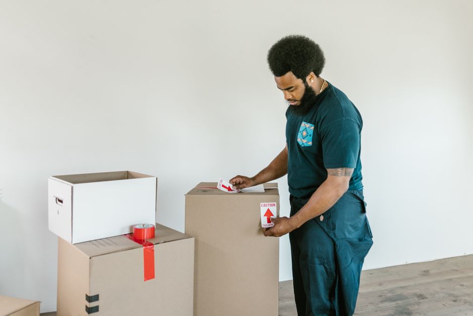 Cardboard moving boxes are stacked neatly in the corner of an empty room with hardwood floors, with bright sunlight streaming through a large window.